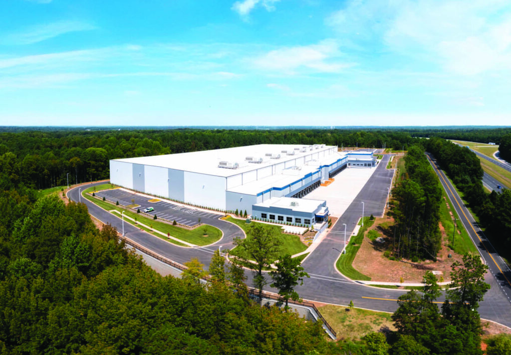 A large white warehouse building surrounded by trees, with multiple loading docks, a few parked trucks, and access roads winding around the facility under a blue sky.