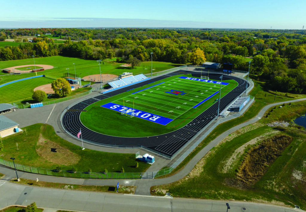 Aerial view of a sports complex featuring a football field with “ROYALS” written in blue end zones, a running track, baseball fields, bleachers, and surrounding trees and greenery under a clear sky.