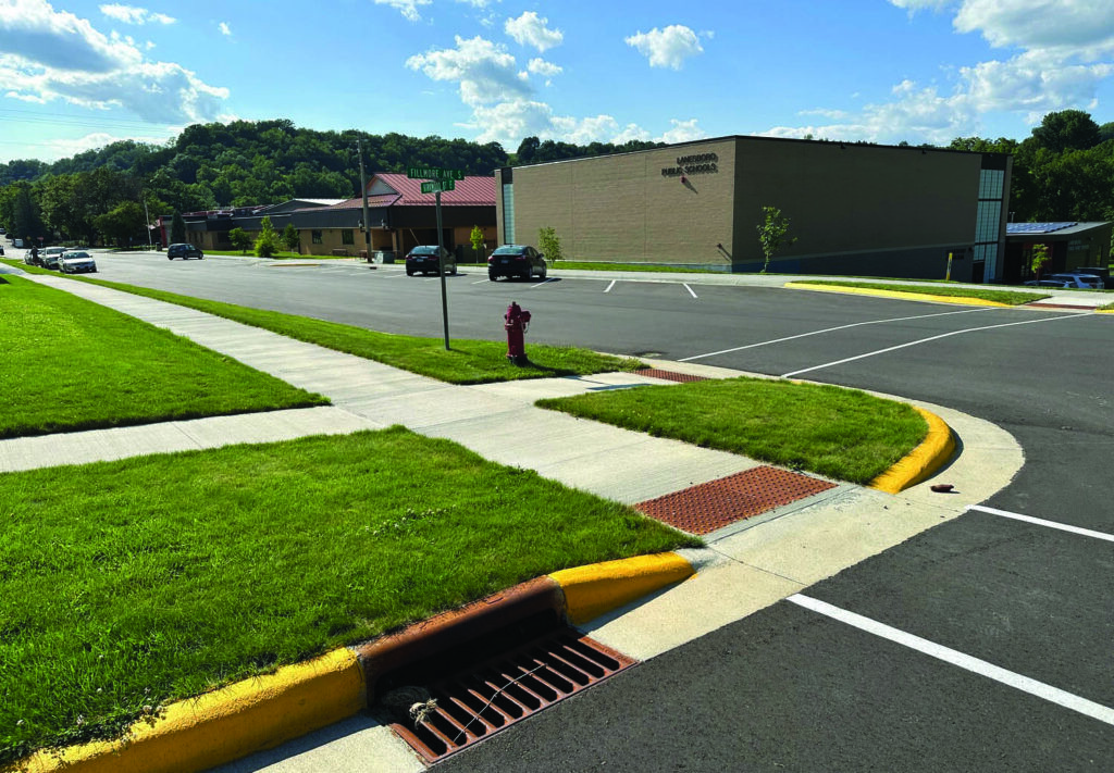 A clean, empty parking lot next to a sidewalk with fresh green grass, a red fire hydrant, and a large tan building in the background under a sunny, blue sky with scattered clouds.