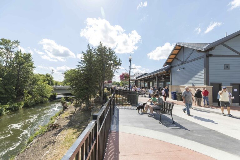 People walk and sit on benches along a riverside pathway next to a modern building under a bright, sunny sky. Trees and a flowing river are on the left, while more people gather near the building on the right.