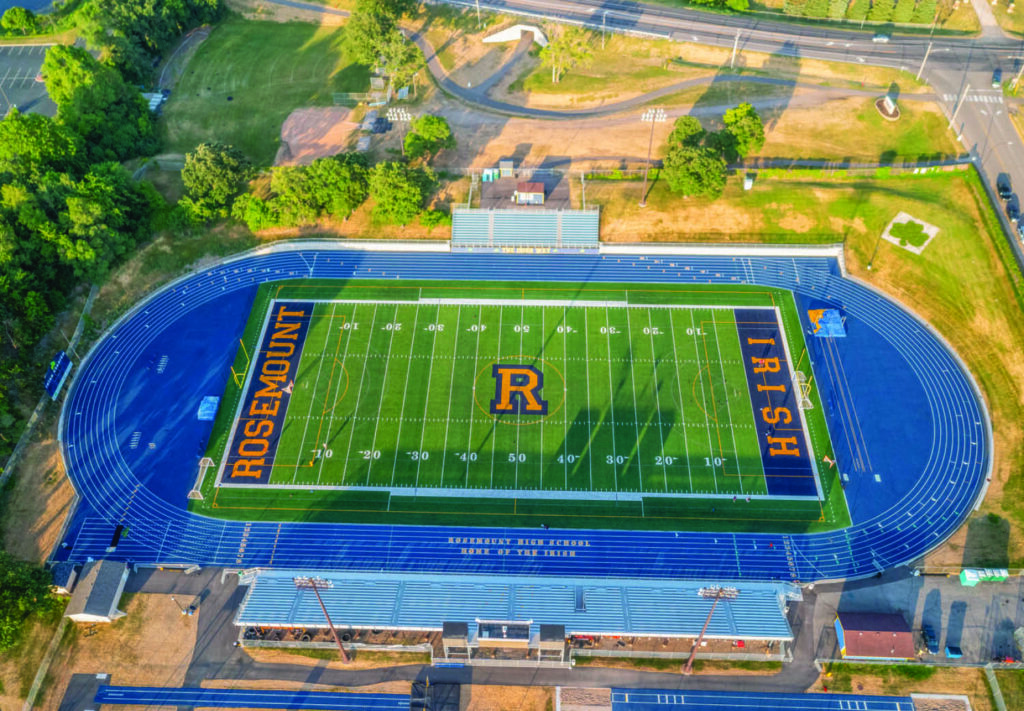 Aerial view of a football field with a blue running track, “Rosemount Irish” written on the field, and a large “R” logo at the center. Bleachers and trees surround the stadium.