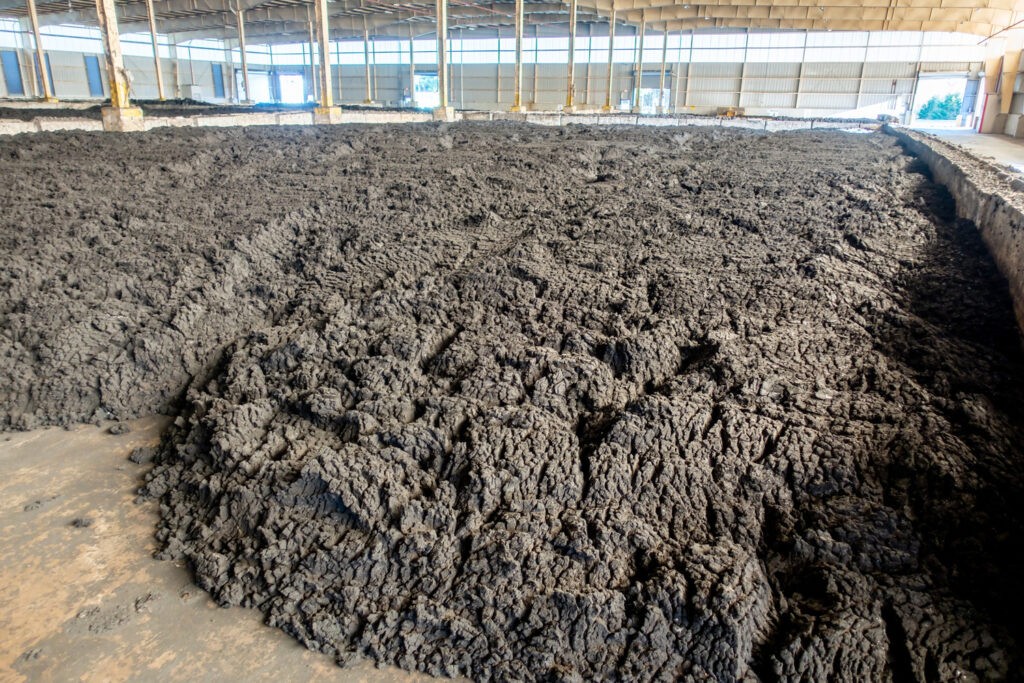 A large indoor facility with a wide, uneven spread of dark, muddy material covering the floor, likely used for composting or waste processing. Structural beams and a partially open wall are visible in the background.