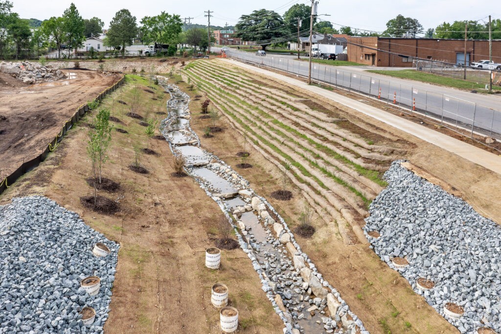 A terraced bioswale with young trees, rows of grasses, and areas of rock lining, runs alongside a road and industrial buildings. The area is under development, with earthmoving visible on one side.