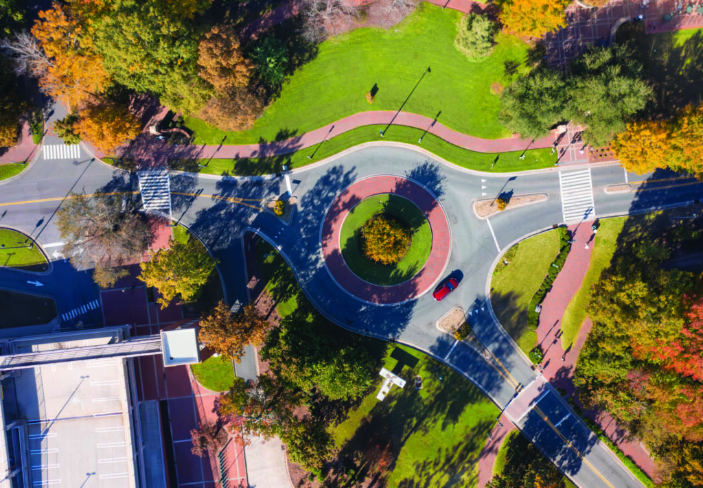 Aerial view of a roundabout with a red car driving through, surrounded by green lawns, trees with autumn foliage, sidewalks, and crosswalks in a park-like urban setting.