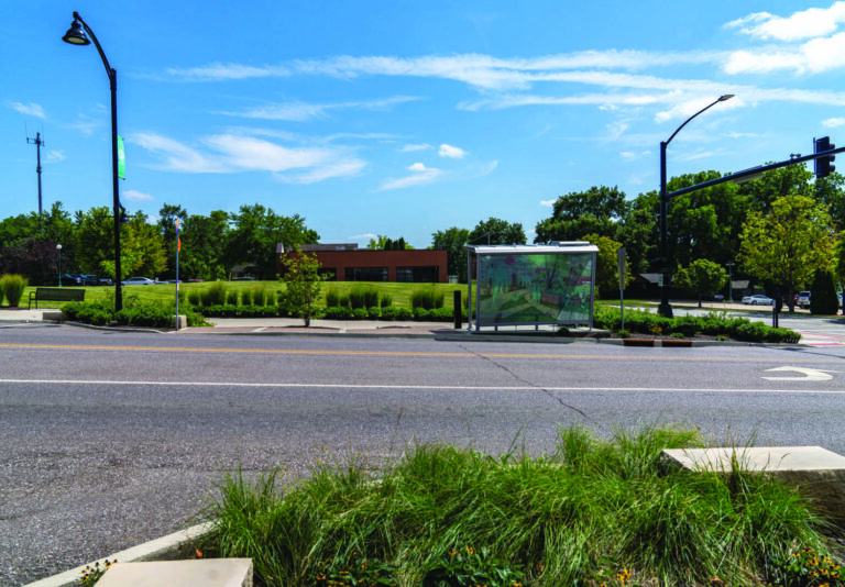 A city street with a modern glass bus stop shelter, green grass and plants in the foreground, trees and a low building in the background, and a blue sky with scattered clouds overhead.
