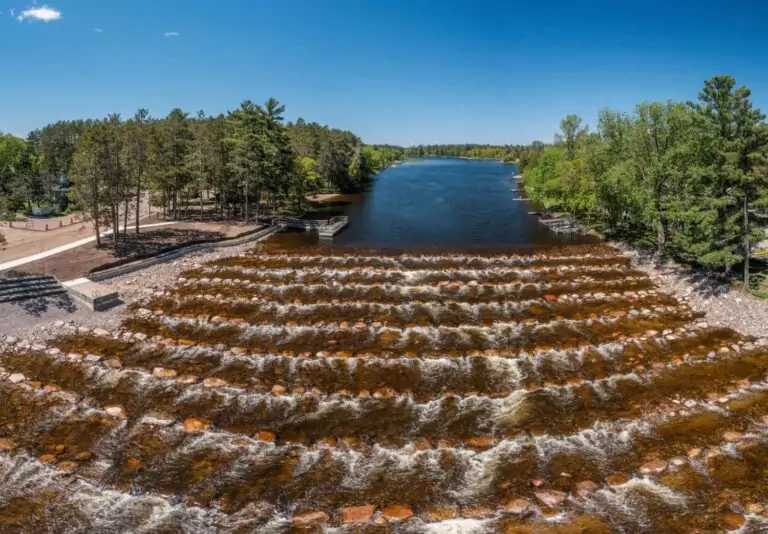 Aerial view of a river with stepped rock formations creating small cascading waterfalls, surrounded by lush green trees and a clear blue sky.