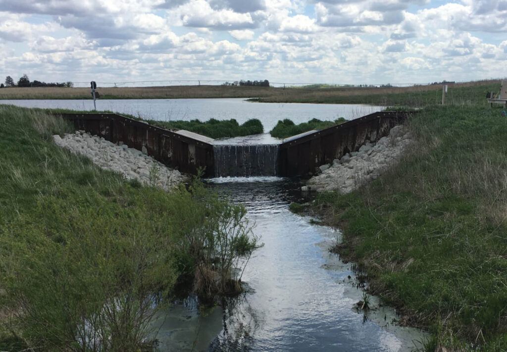 A small dam with water flowing over its edge into a narrow stream, surrounded by grassy banks and rocks, under a cloudy sky in a rural landscape.