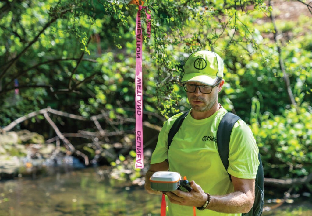 A person wearing a neon yellow shirt and cap stands outdoors by a stream, holding a device. Pink survey tape hangs from a tree branch above, and the background is lush with green foliage.