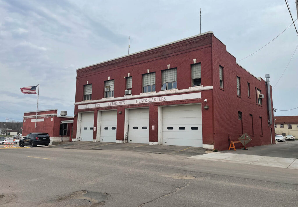 A large red brick fire department headquarters building with three garage doors, several windows, and an American flag flying outside on a cloudy day.