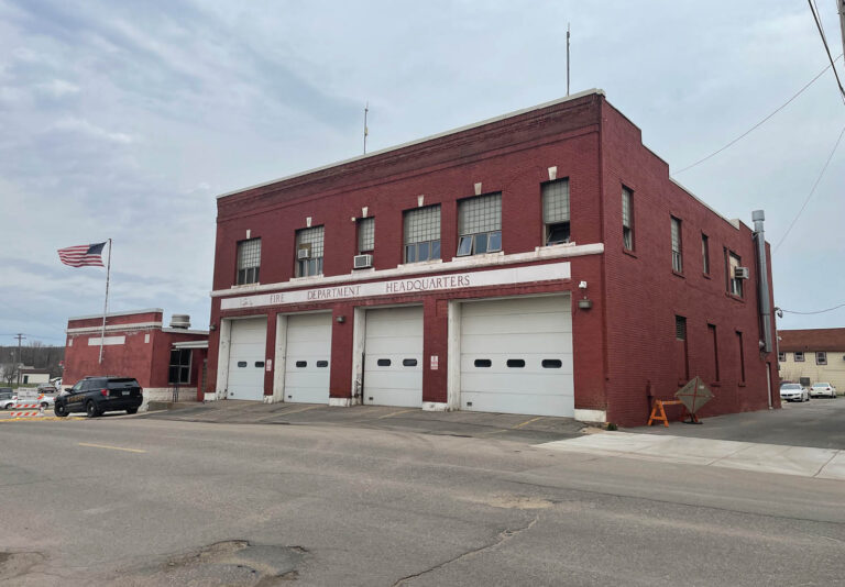 A large red brick fire department headquarters building with three garage doors, several windows, and an American flag flying outside on a cloudy day.