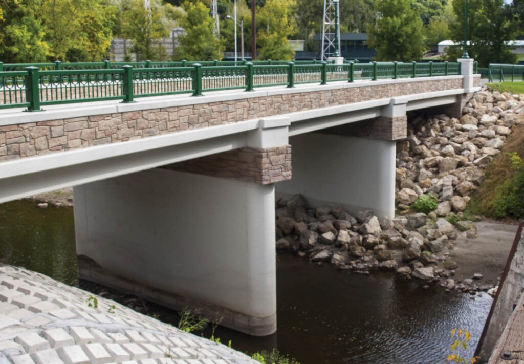 A concrete and stone bridge with green railings spans over a shallow river with rocks along the banks. Trees and grass are visible in the background.
