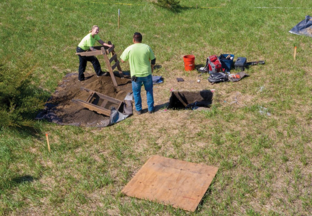 Two people in green shirts conduct an archaeological dig on grass, using tools and sifting soil through a screen. Excavated pits, equipment, and a wooden board are visible around them.