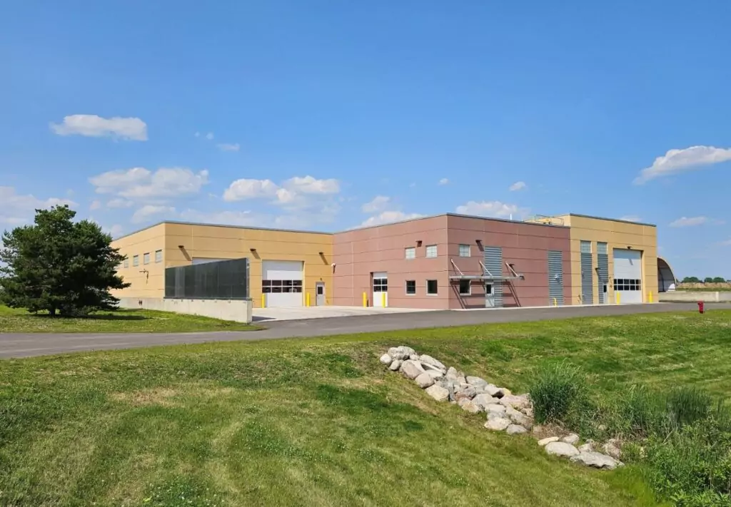 Modern industrial building with beige and red exterior walls, multiple large garage doors—some open—and smaller windows, set along a paved driveway with grassy surroundings, rocks, and shrubs under a partly cloudy sky.