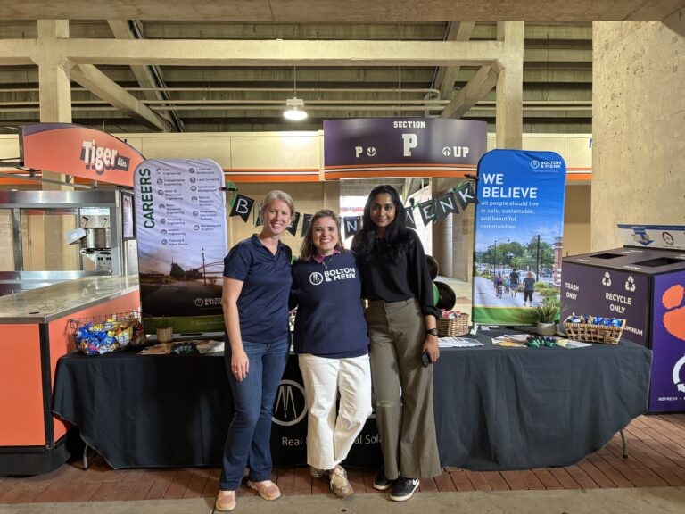 Three women stand smiling in front of a career fair booth with banners and promotional materials displayed on the table, proudly representing Empowering Communities.