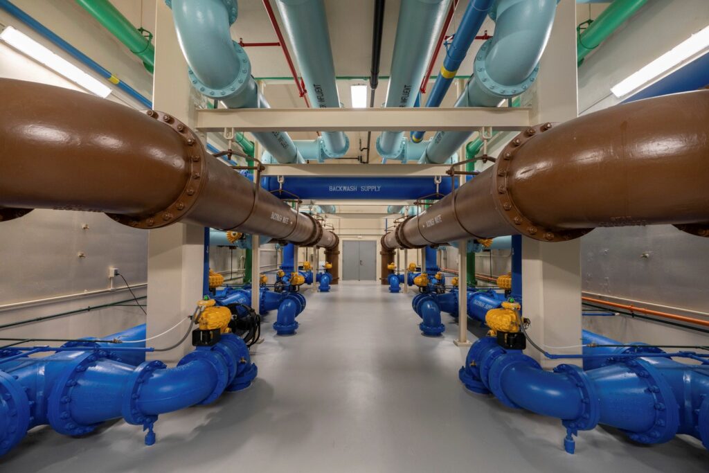 A symmetrical industrial room features large brown, blue, and turquoise pipes running parallel along the ceiling and floor—showcasing a clean, CMAR Project Delivery approach. Yellow valve mechanisms and a sign reading “BACKWASH SUPPLY” complete the space.