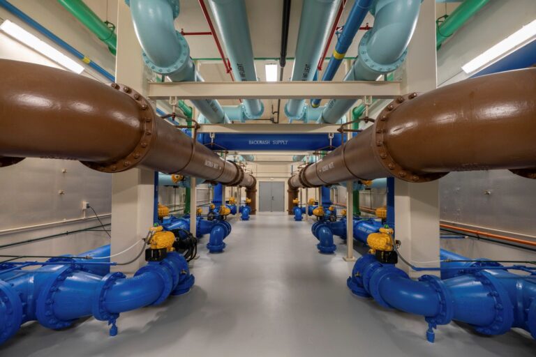 A symmetrical industrial room features large brown, blue, and turquoise pipes running parallel along the ceiling and floor—showcasing a clean, CMAR Project Delivery approach. Yellow valve mechanisms and a sign reading “BACKWASH SUPPLY” complete the space.