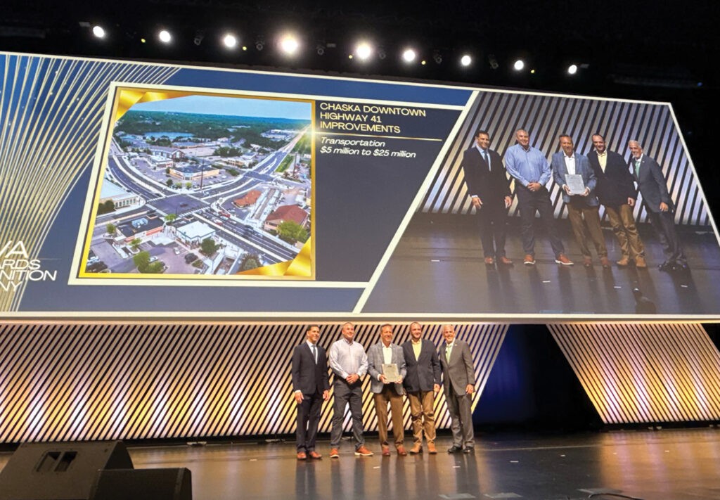A group of six people stand on stage receiving an award. Behind them, a large screen displays an aerial image of the City of Chaska and text about the Downtown Highway 41 Improvements project, with a cost range of $5 to $25 million.