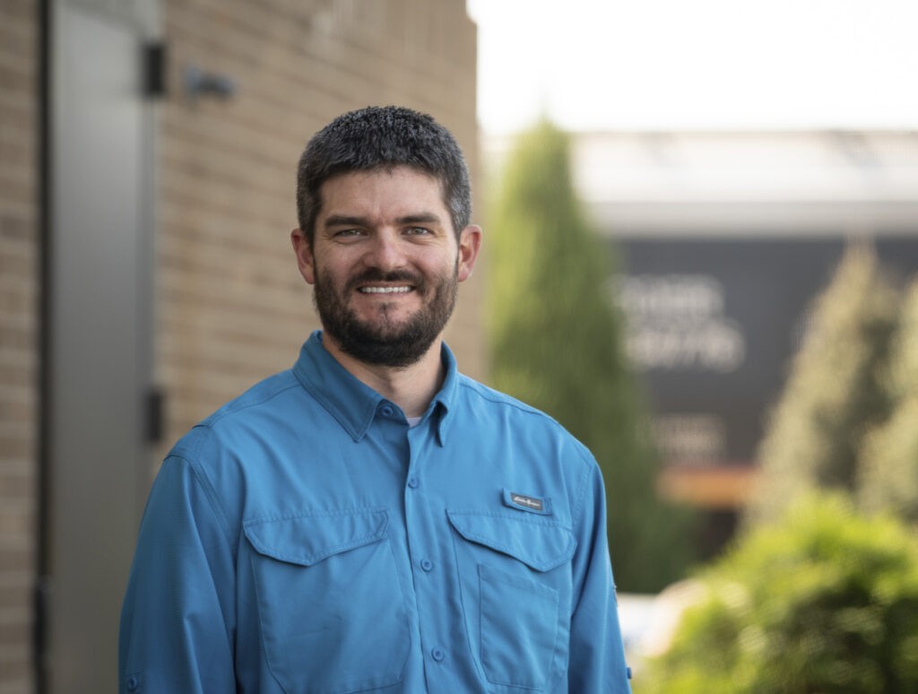 A man with short dark hair and a beard, wearing a blue button-up shirt, stands smiling outdoors next to a brick building with greenery.