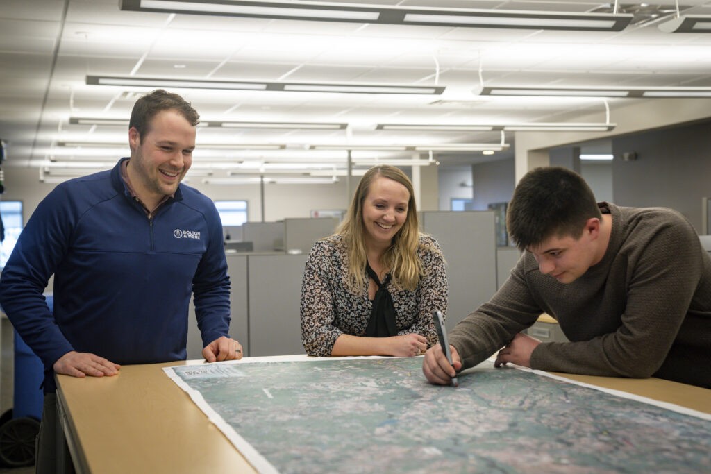 Three people stand around a large map on a table in an office. One person is marking the map with a pen while the other two look on and smile. The workspace is modern with overhead lighting and cubicles.