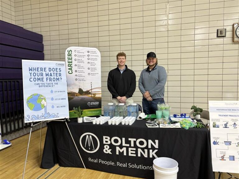 Two men stand behind a Bolton & Menk display table, empowering communities with water bottles, plants, and educational materials about water sources in a gymnasium setting with informational posters behind them.