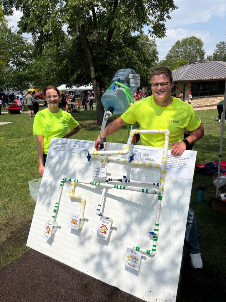 Two people in bright yellow shirts stand outdoors beside a water system display with pipes and labels, empowering communities by demonstrating how it works. One pours water from a jug as people and tents fill the sunny background.