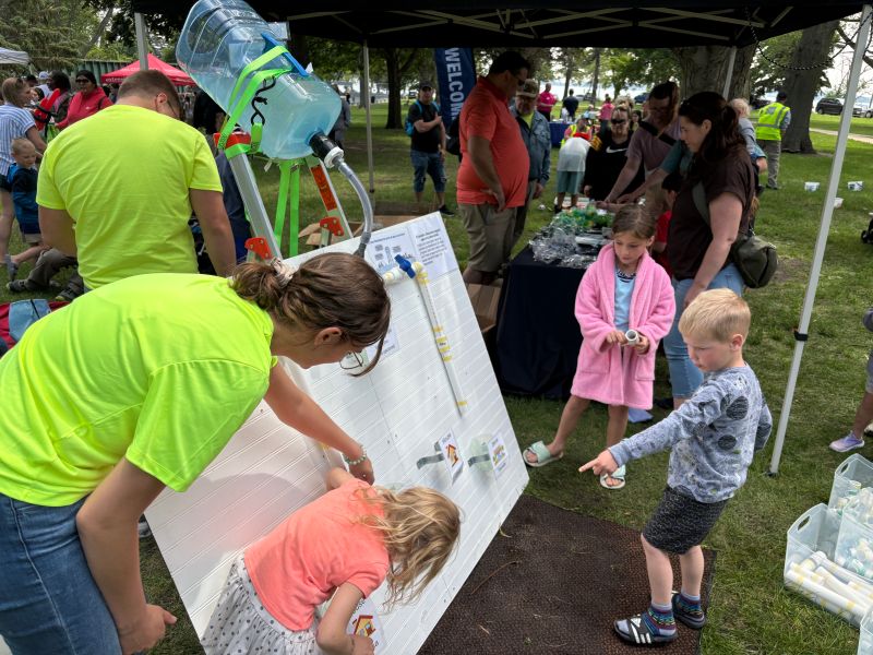 Children and adults gather outdoors around a hands-on science activity with water flowing through tubes on a large vertical board, empowering communities to explore STEM together; tables and other event attendees are visible in the background.