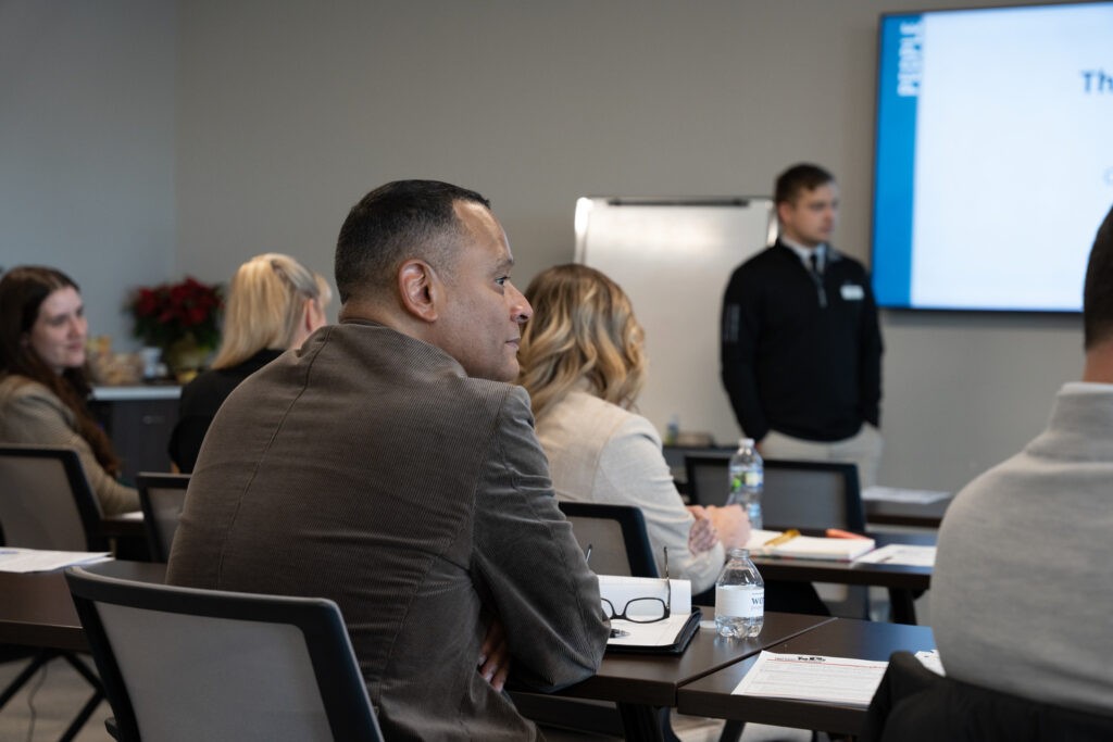 People seated at tables in a classroom, attentively listening to a presenter standing by a screen and whiteboard at the front of the room. Notebooks, papers, and water bottles are on the tables.