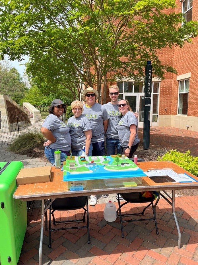 Five people stand behind an outdoor table with a colorful display model and informational materials, empowering communities. They are smiling in matching gray shirts, with trees and a brick building visible in the background.