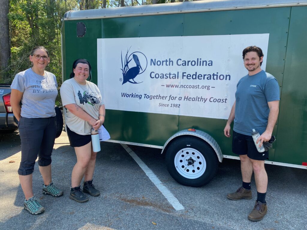 Three people stand smiling in front of a green trailer with a sign reading North Carolina Coastal Federation: Working Together for a Healthy Coast, highlighting their role in empowering communities during an outdoor event.