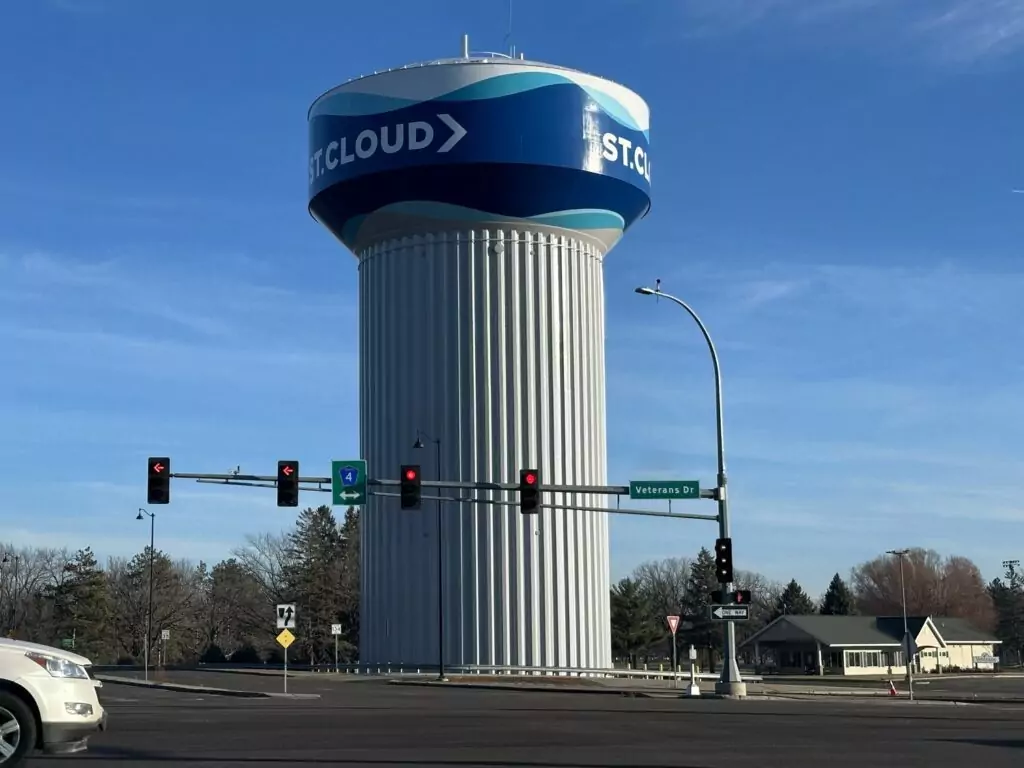 A tall, cylindrical water tower labeled ST. CLOUD stands behind a street intersection with traffic lights and signs, including one for Veterans Drive. Trees and houses are visible in the background.