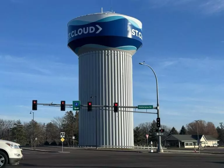 A tall, cylindrical water tower labeled ST. CLOUD stands behind a street intersection with traffic lights and signs, including one for Veterans Drive. Trees and houses are visible in the background.