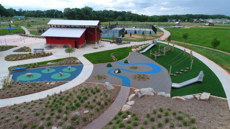 Aerial view of a modern park with a red barn-style building, tennis courts, green lawns, a childrens playground with slides, climbing structures, and spinning equipment, surrounded by walking paths and landscaped areas.