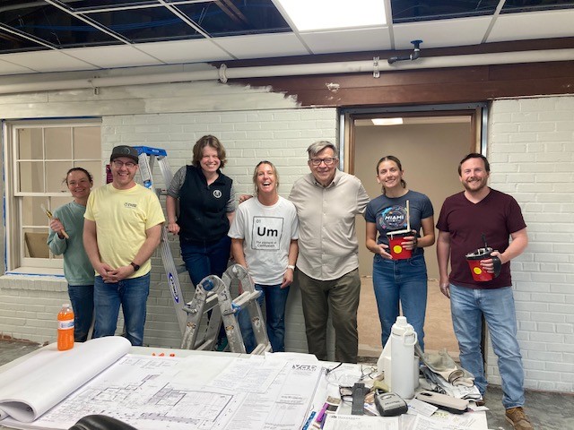 Seven people stand smiling in a partially renovated room with exposed ceiling and ladders, holding painting supplies. Blueprints, drinks, and painting tools are on a table in the foreground.