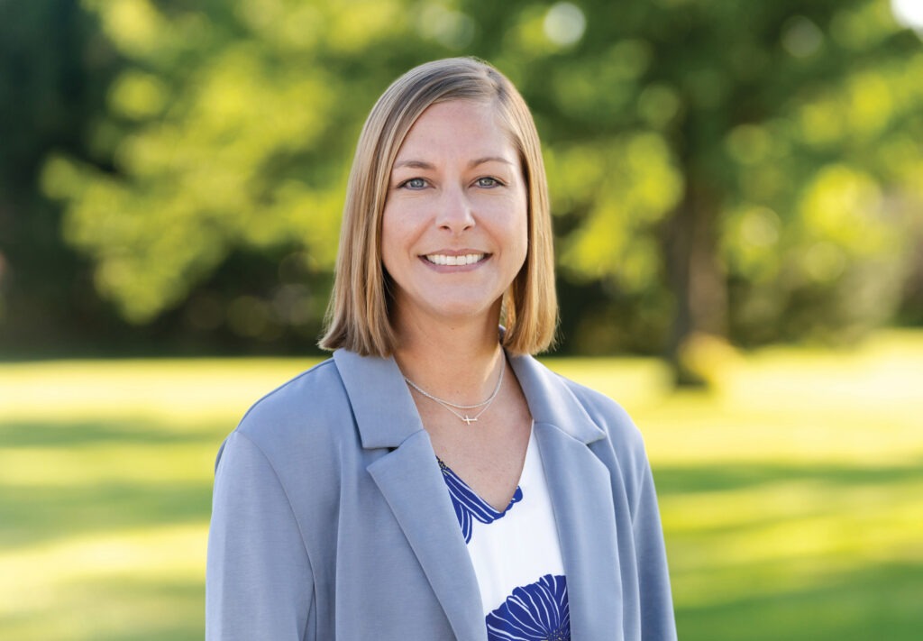 Person with short light brown hair wearing a light gray blazer over a white blouse with blue floral patterns, standing outdoors in a park-like setting with green grass and trees in the background.