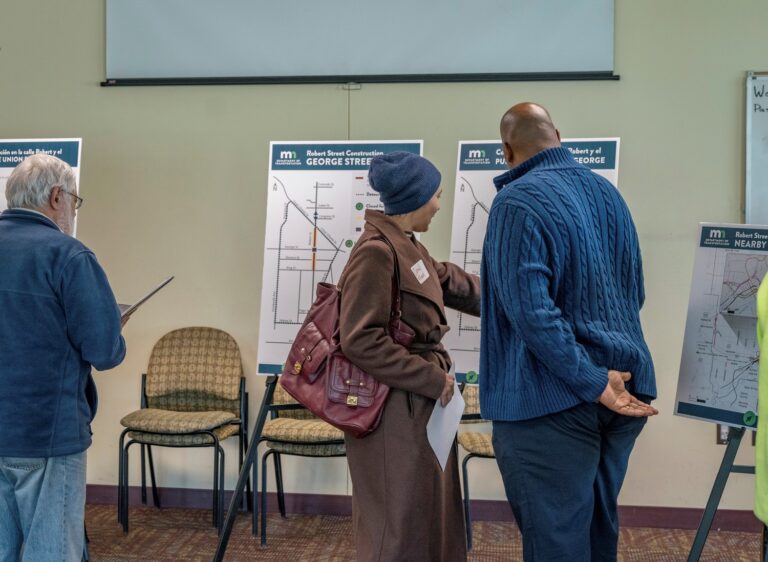 Three people stand in front of informational posters displaying street maps at a community meeting. One woman points to a map while talking to a man; another man reads a paper nearby.