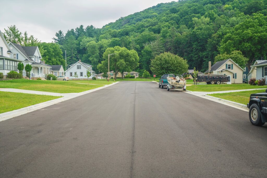 A quiet suburban street with houses on both sides, green lawns, parked cars, and a lush, tree-covered hill in the background under a cloudy sky.