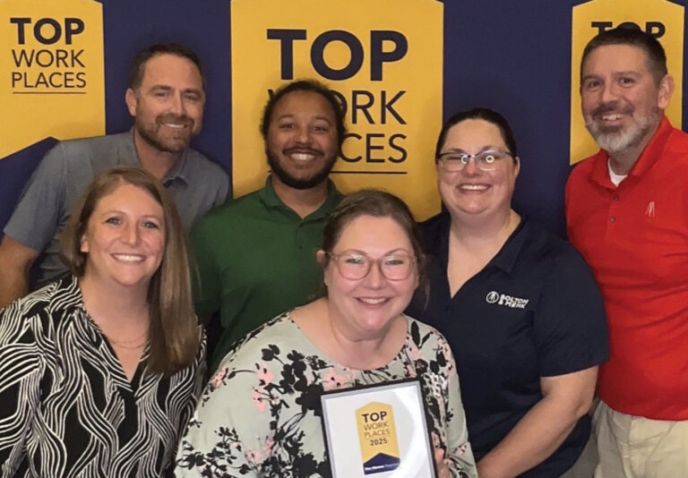 Six smiling people pose together in front of a “Top Work Places” backdrop, with one person holding a plaque that reads “Top Work Places 2023.”.