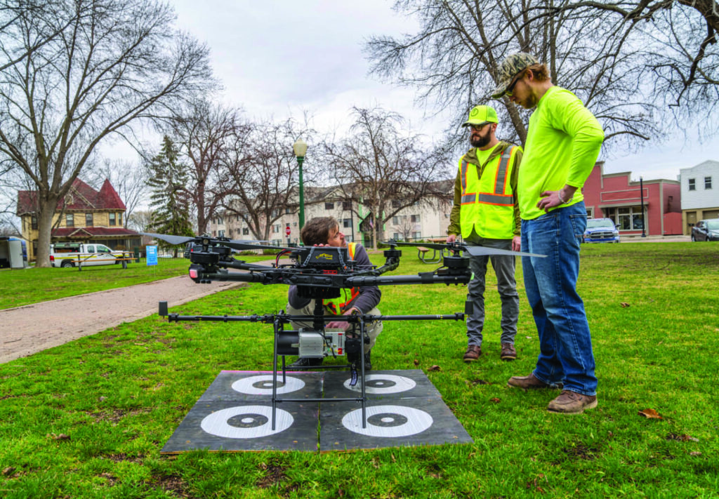 Three workers in safety gear stand in a park while one adjusts a large drone on a landing pad with target symbols. Trees and buildings are visible in the background.