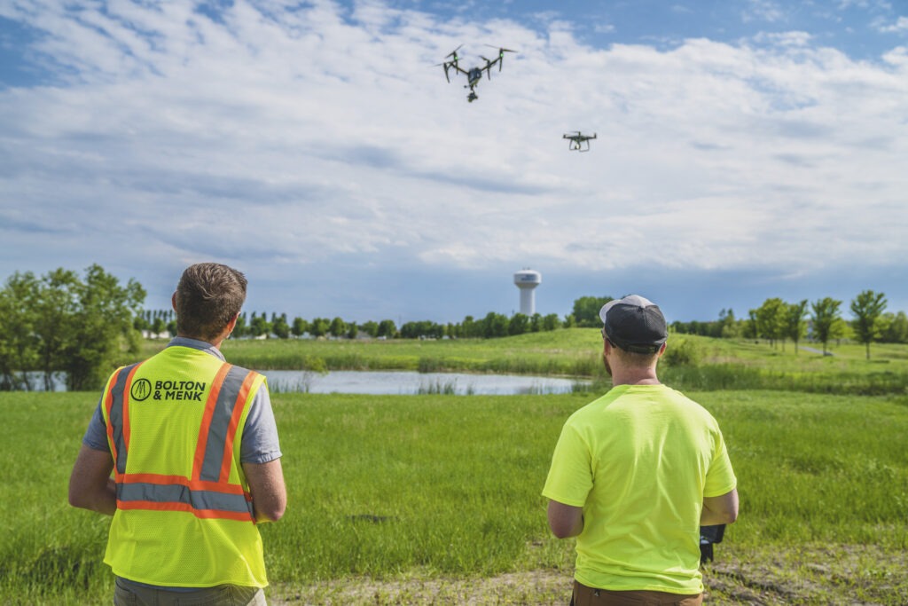 Two people in safety vests operate drones over a grassy field near a pond, with a water tower and trees visible in the background under a partly cloudy sky.
