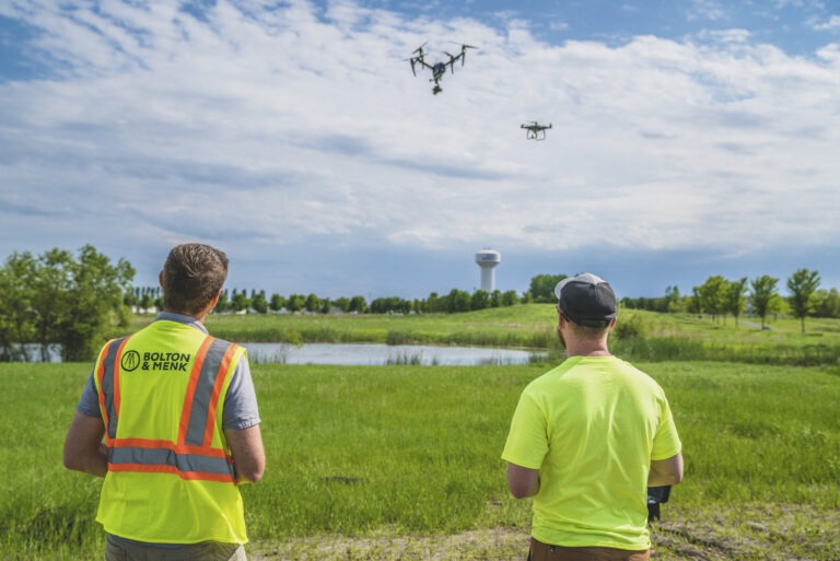 Two people in safety vests operate drones over a grassy field near a pond, with a water tower and trees visible in the background under a partly cloudy sky.