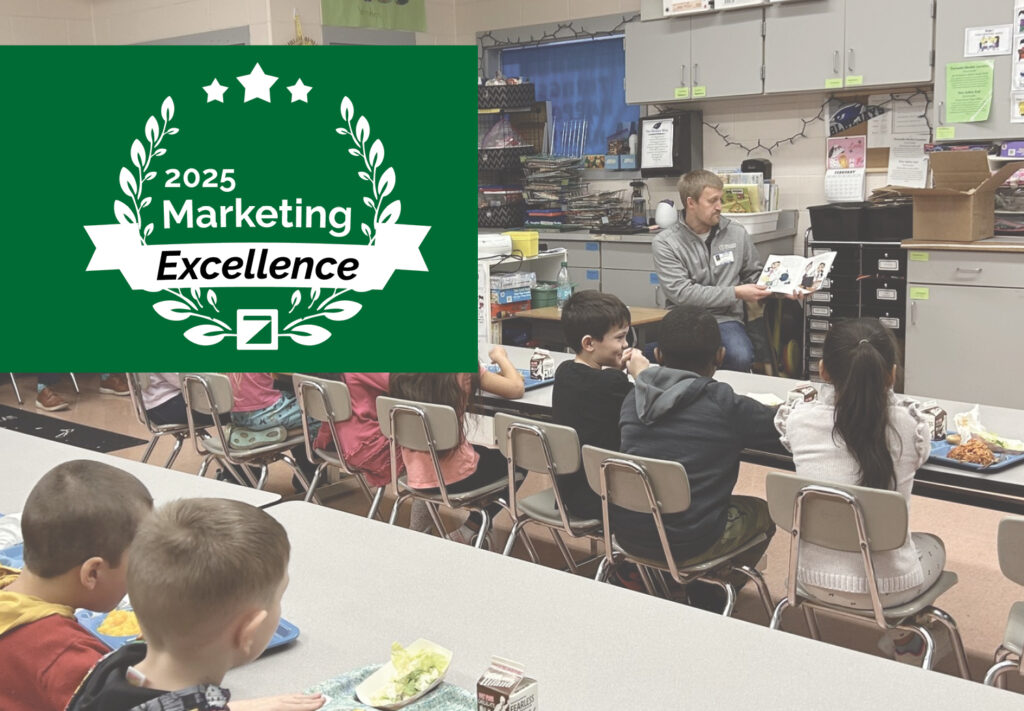 A man reads a book to a group of young children seated at tables in a classroom. Overlaid is a green 2025 Marketing Excellence award graphic featuring laurel leaves and stars.