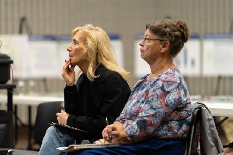 Two women sit side by side, attentively listening. One holds a pen to her chin and a clipboard, while the other holds a notepad.