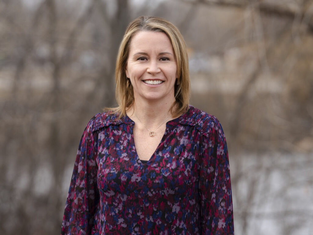 A woman with shoulder-length blonde hair is smiling outdoors. She is wearing a patterned blouse in shades of purple and blue. The background is blurred, suggesting bare trees and a muted landscape, reflecting her passion for PFAS Solutions in natural environments.