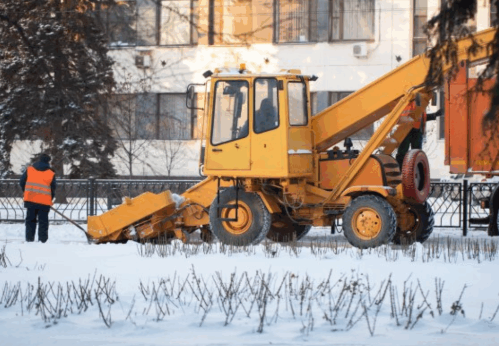 A yellow snow removal vehicle clears snow from a street near a building while a worker in an orange vest stands nearby. Snow covers the ground and leafless shrubs are visible in the foreground.