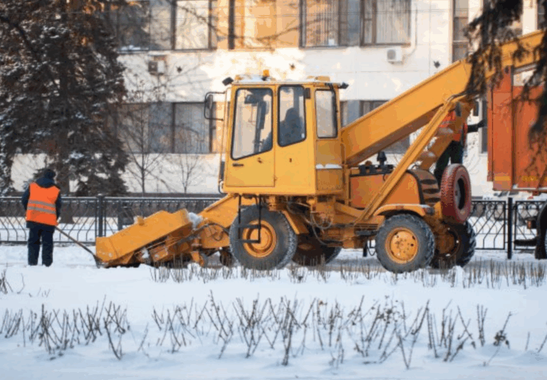 A yellow snow removal vehicle clears snow from a street near a building while a worker in an orange vest stands nearby. Snow covers the ground and leafless shrubs are visible in the foreground.