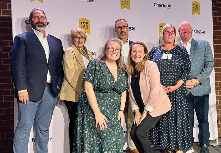 A group of seven professionally dressed adults poses and smiles together in front of a step-and-repeat backdrop featuring Top Workplaces and Charlotte logos.