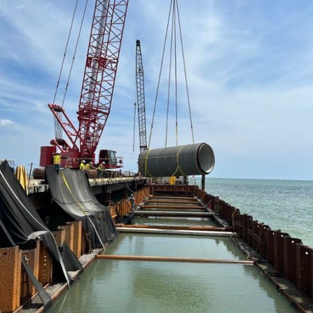 Large cylindrical steel pipe being lifted by a red crane on a pier construction site over water, with another crane in the background and calm blue sky overhead.
