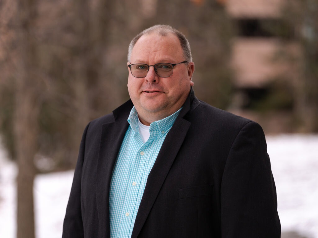 A middle-aged man wearing glasses, a black blazer, and a light blue checkered shirt stands outdoors on a snowy day, embodying professionalism often found in asset management, with blurred trees and buildings in the background.