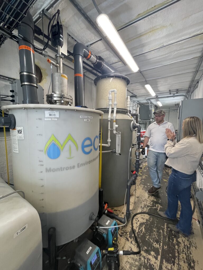 Two people stand inside an industrial facility next to large tanks labeled “eco Montrose Environmental,” discussing PFAS Solutions and equipment, including pipes and gauges, in a well-lit room.
