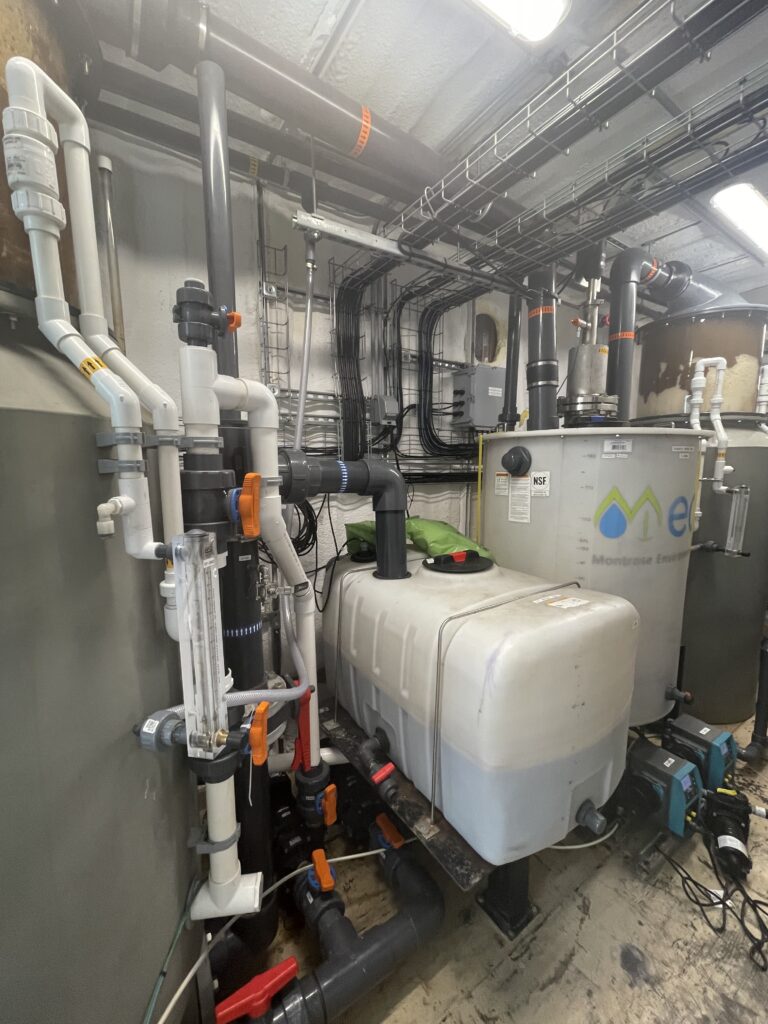 A mechanical room with large white and gray tanks, various pipes, valves, meters, and electrical wiring on the walls and ceiling.